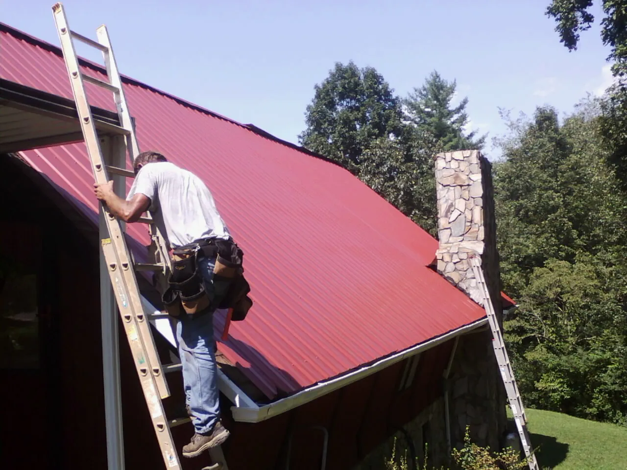 Roofer on a ladder beside a red metal roof and stone chimney — Soffit & Fascia Repair by Roofing Brothers in Dundas
